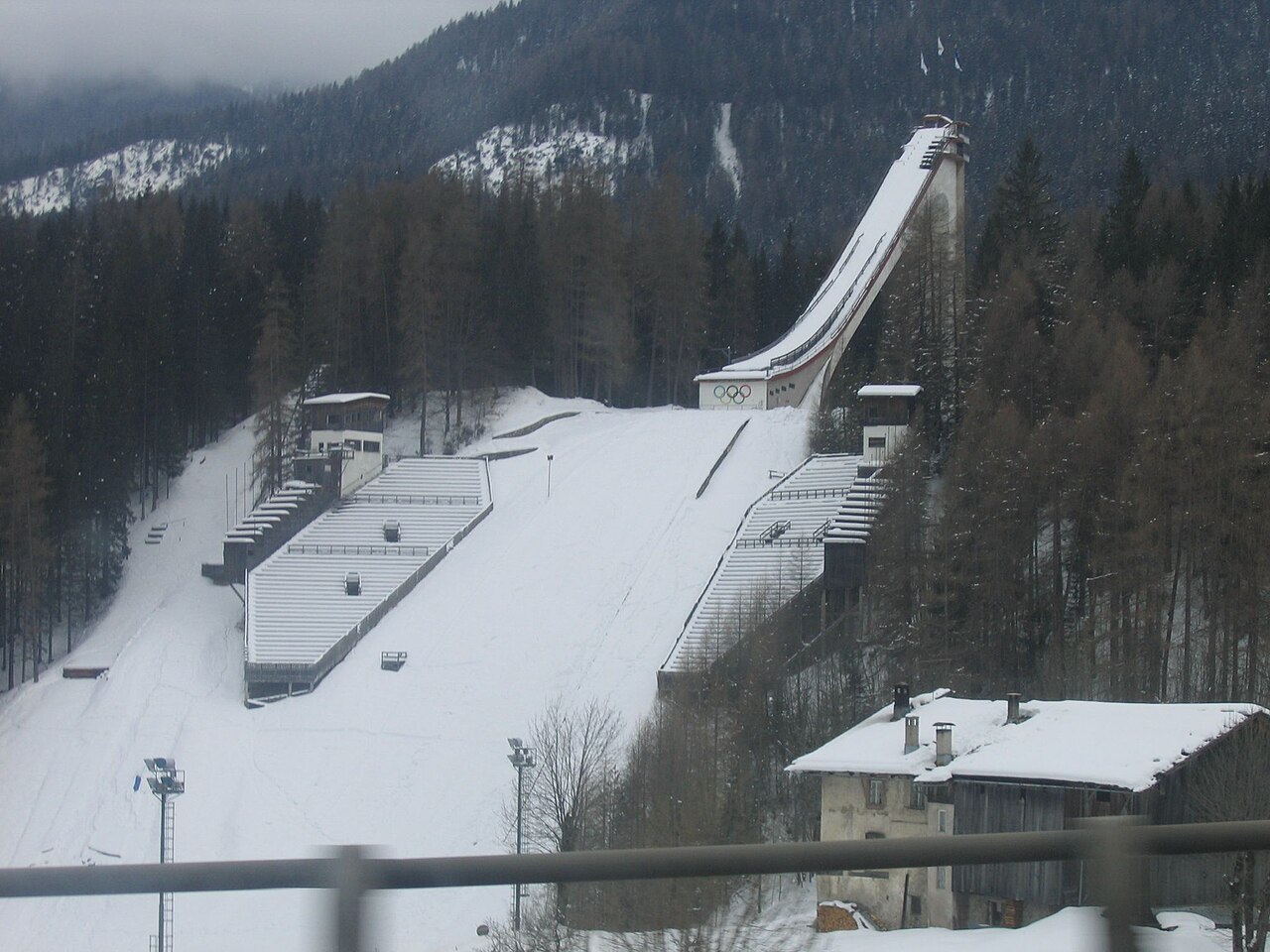 Snow covers the historic Trampolino Olimpico ski jump in Cortina d’Ampezzo, with wooded mountains behind it.