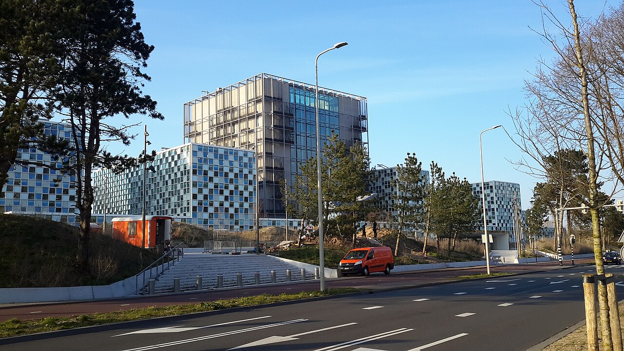 International Criminal Court headquarters in The Hague, seen across a road with trees and a red van in front.