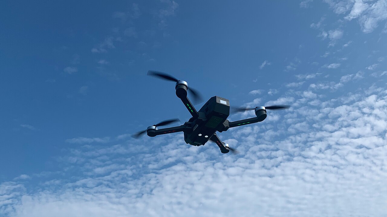 A quadcopter drone seen from below in flight against a blue sky.