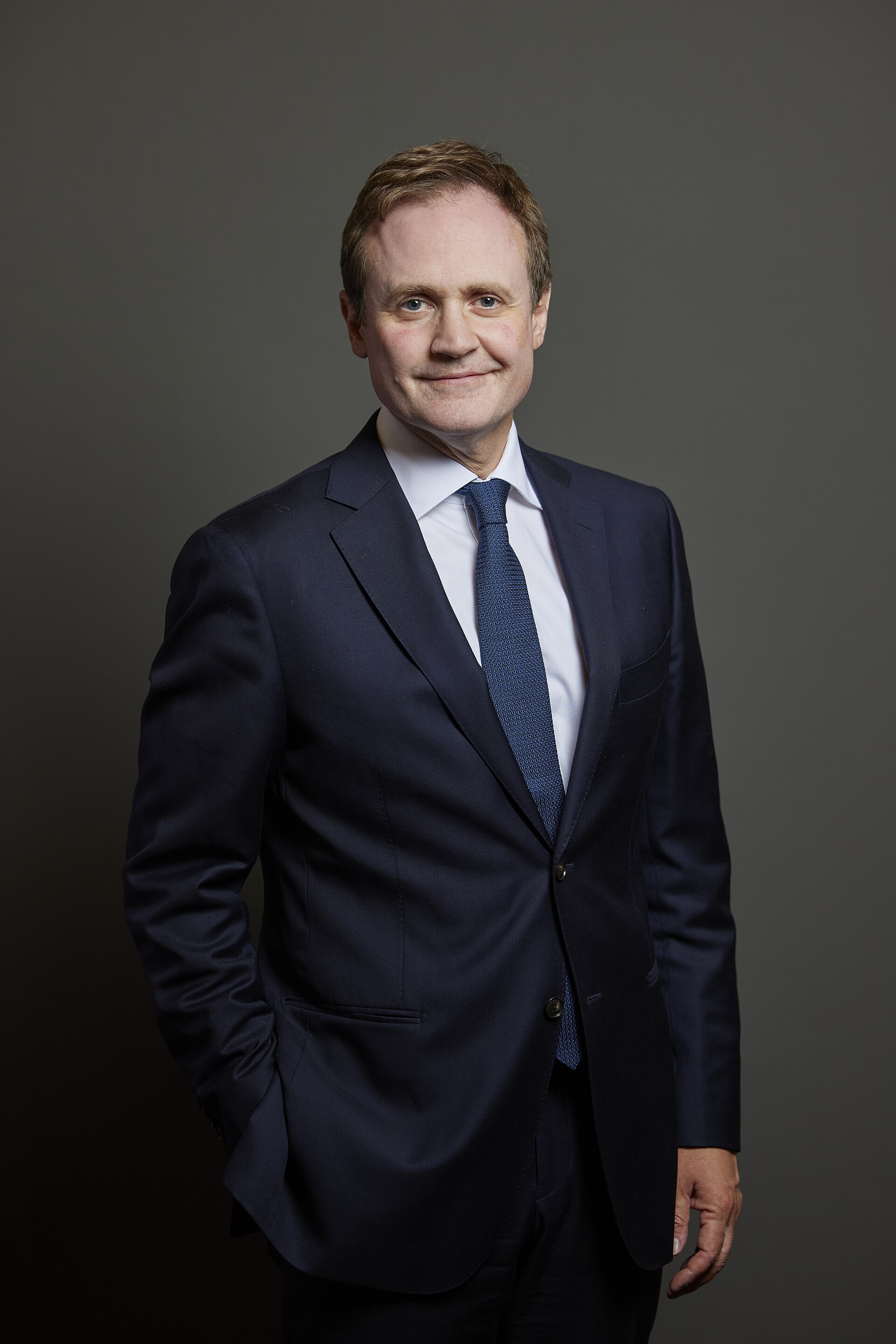 Official portrait of Tom Tugendhat in a dark suit and blue tie against a gray background.