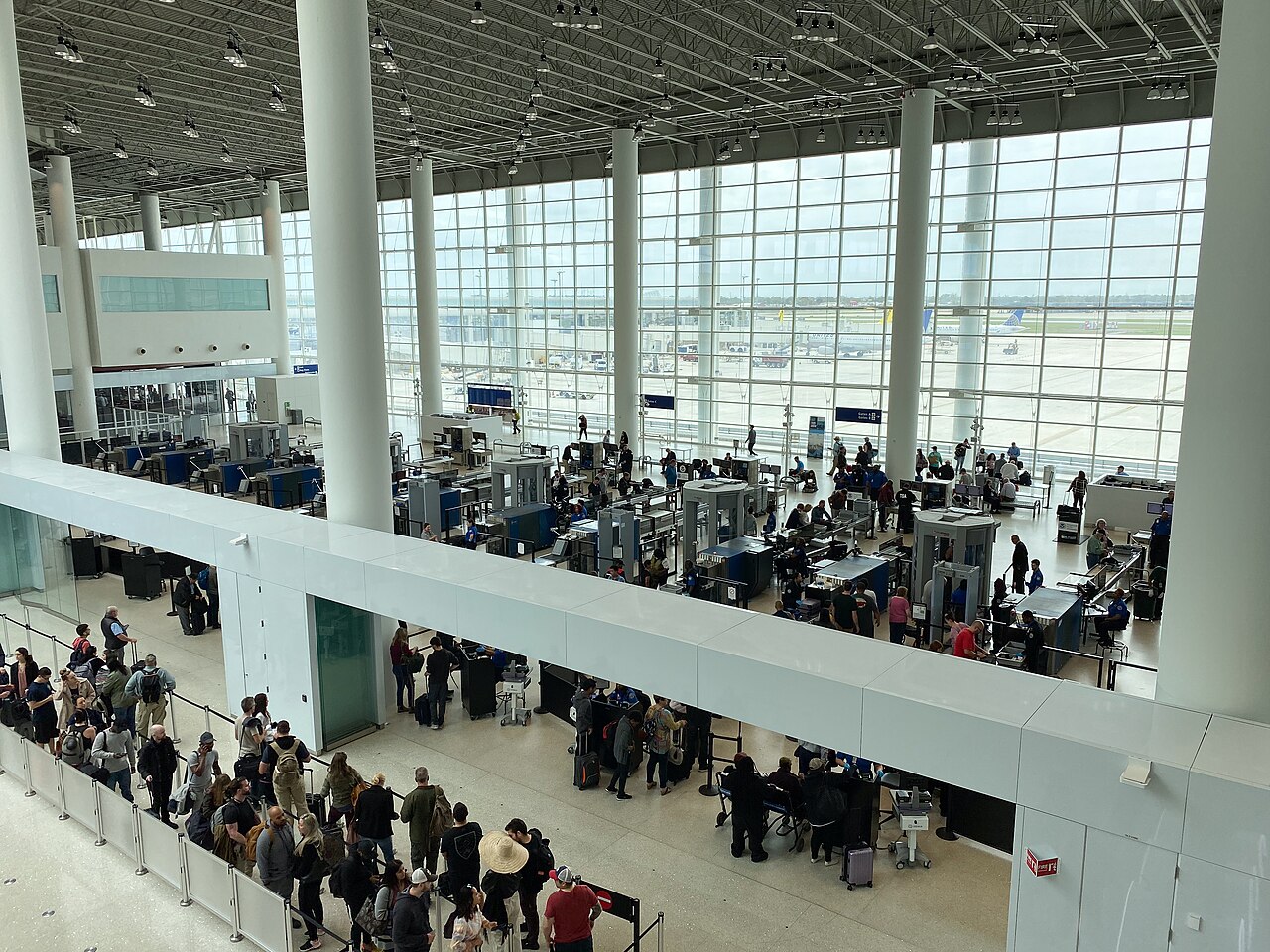 Passengers queue beneath large terminal windows at a TSA security checkpoint in New Orleans airport.