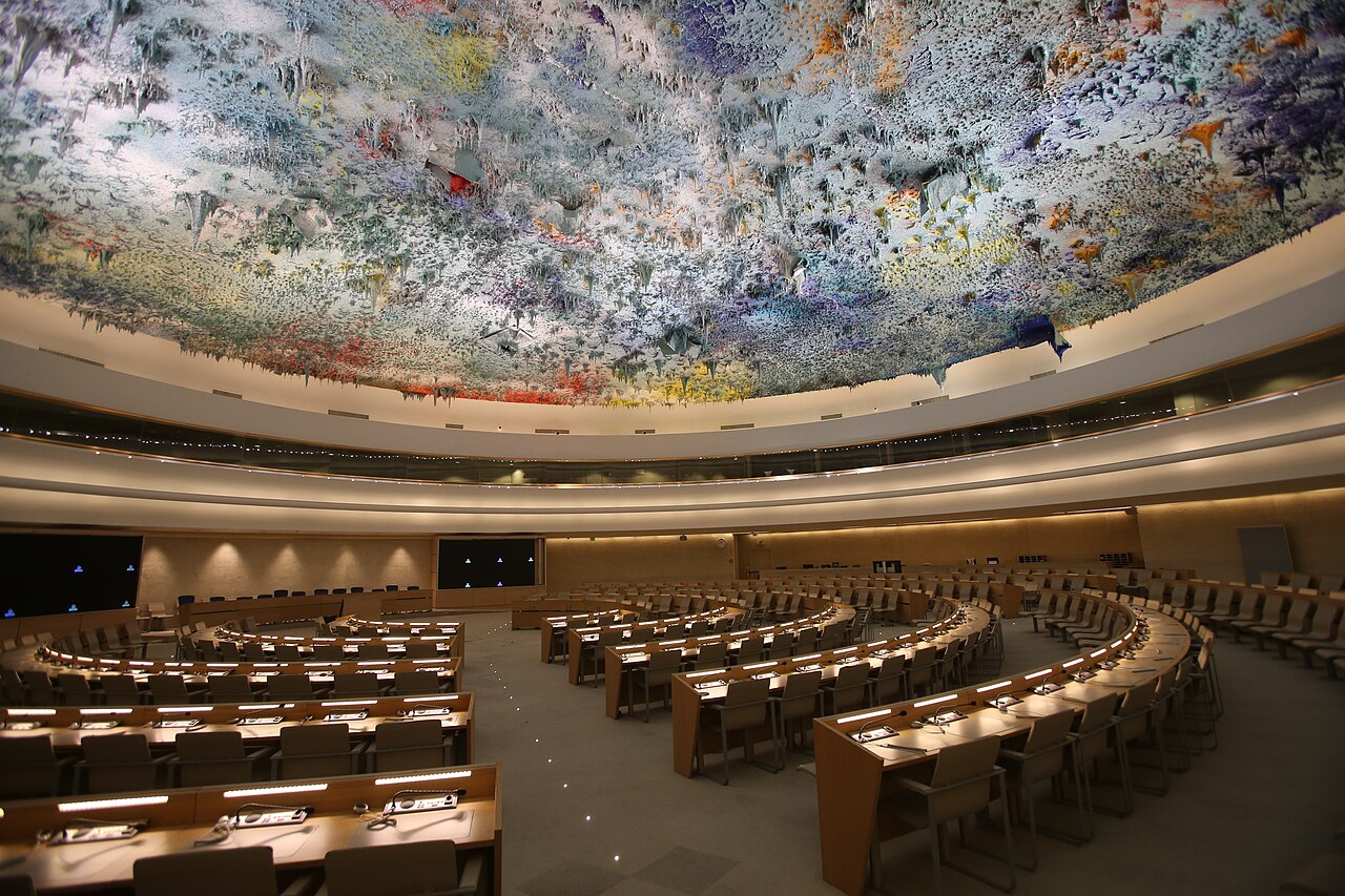 Empty curved rows of seats beneath the colorful ceiling of the UN Human Rights Council meeting room in Geneva.